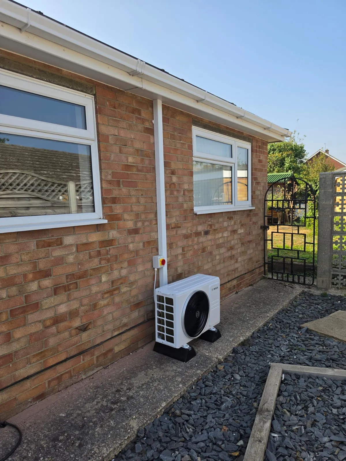 Wider view — condenser unit on a path along the side of the house, white trunking running up the wall